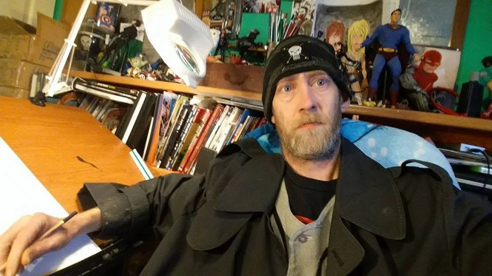 A close selfie of the artist at his drawing desk, with action figures and comics on the shelf behind him.