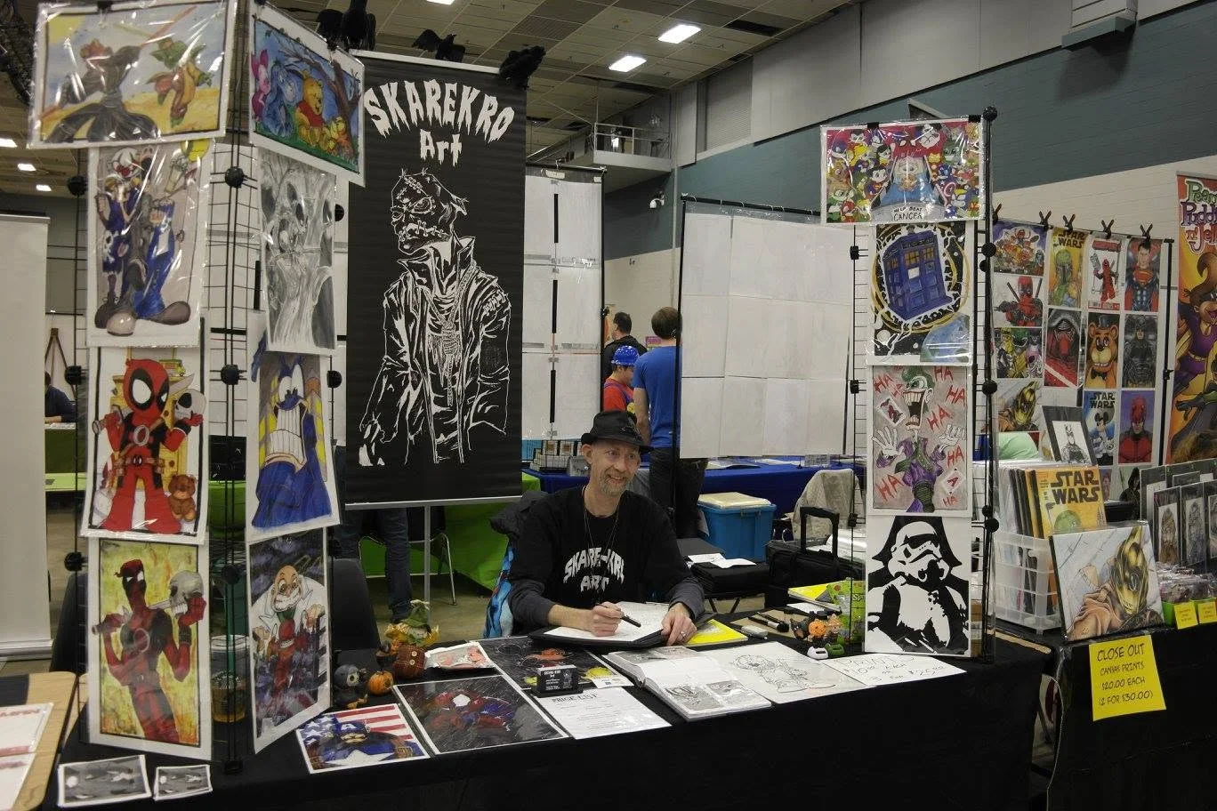 The artist smiling at his convention table, wearing a Skarekro Art shirt and hat, with comics and prints displayed behind him.